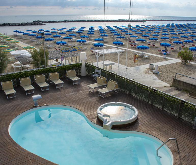 Spiaggia con ombrelloni blu, piscina e jacuzzi su terrazza in legno.